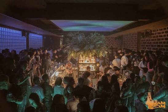 BAM BAR 高峰时段：吧台四周站满客人，顶部投下青绿色光，垂挂绿植装置在中央