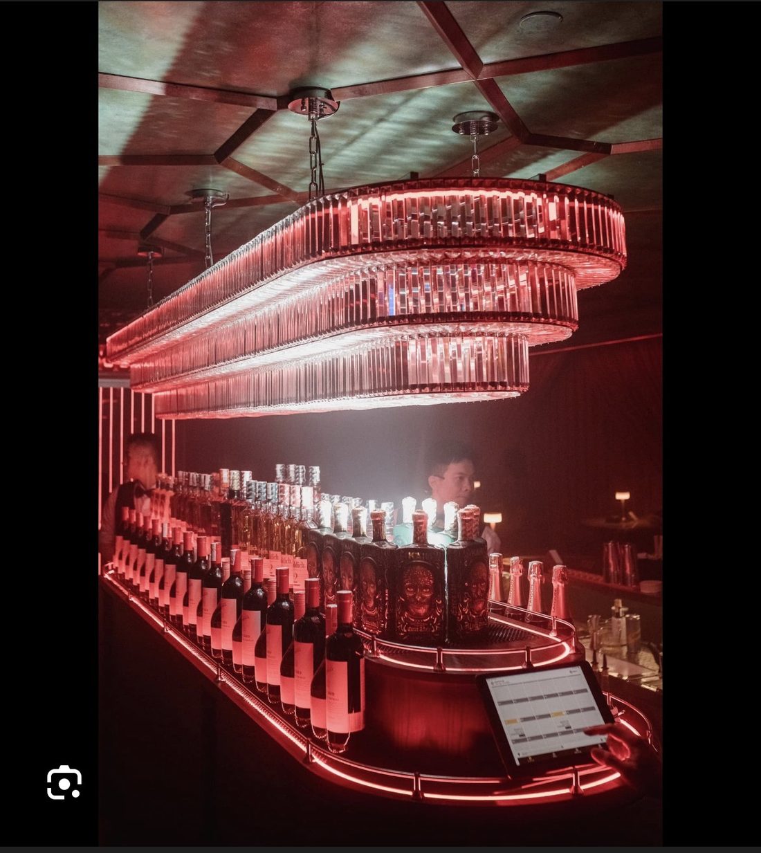 BODEGA SAIGON 吧台：三层长条水晶吊灯下的整排洋酒与气泡酒，红色灯带勾出弧形台沿