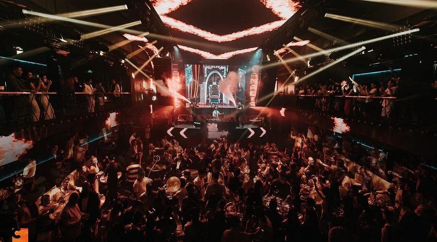 CANALIS CLUB 胡志明夜店主舞池 · 天花板环形火焰灯带、中央 LED 主舞台与两侧二层看台，全场站满客人