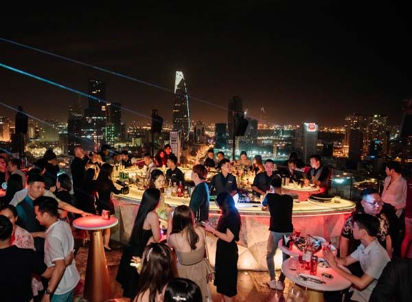 CHILL SKY BAR 天台人群围着吧台，远处能看到 Bitexco 金融塔尖顶与西贡市区夜景