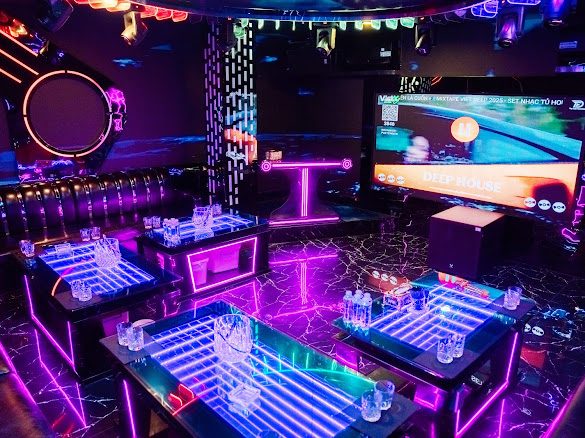 点歌大屏包厢：壁挂大屏正放越南语 Deep House 歌单，中间抬高的台阶用粉色灯带勾边，蓝紫发光茶几